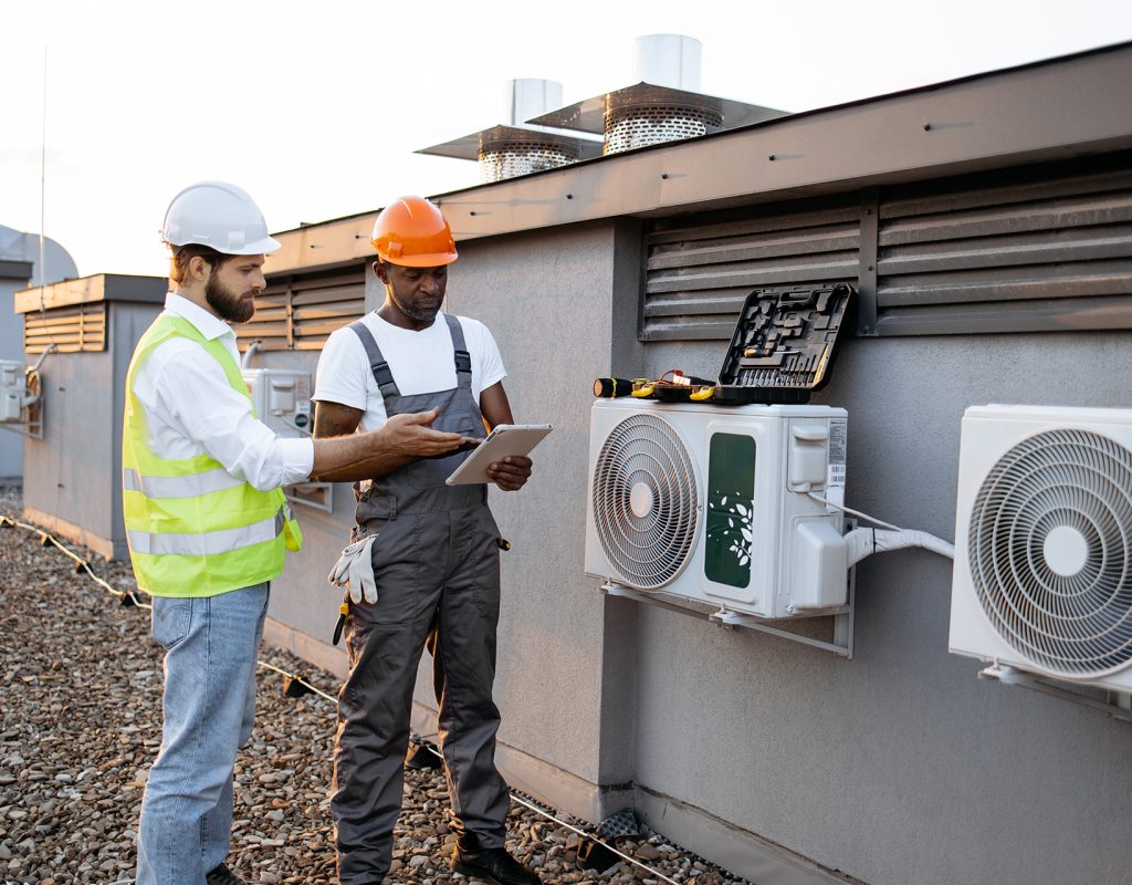 Multicultural males advising with each other about repairing of air conditioner while standing near broken cooling system outdoors. Men using tablet for scrolling websites with useful information.